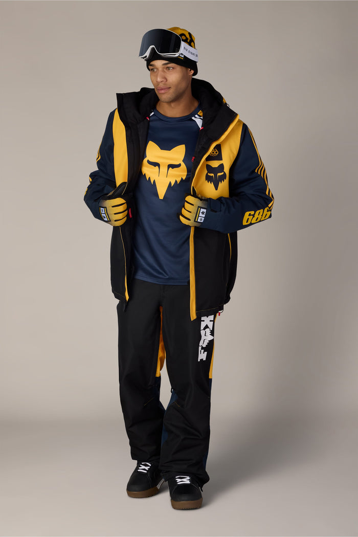 A man wearing a Fox Racing beanie and goggles, a navy and yellow snowboarding jacket, and 686 Men’s Fox Racing Spec Shell Pant.