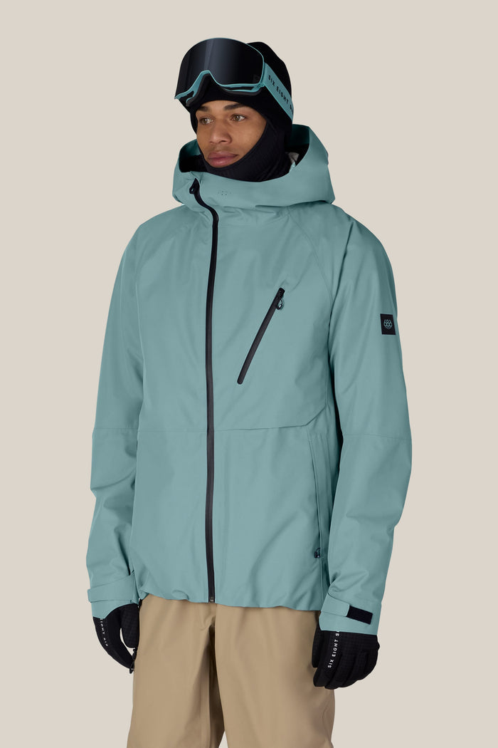 Wearing the 686 Men’s Hydra™ Thermagraph® Jacket in teal by 686, along with black gloves, tan pants, ski goggles, and a black balaclava, this person stands against a plain beige background.