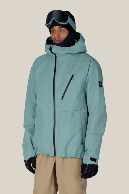 Wearing the 686 Men’s Hydra™ Thermagraph® Jacket in teal by 686, along with black gloves, tan pants, ski goggles, and a black balaclava, this person stands against a plain beige background.