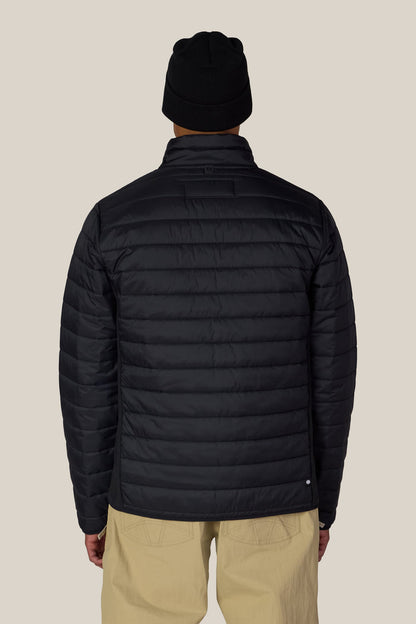 A person is seen from behind, standing against a plain light background, wearing tan pants and the 686 Men’s Thermal Puff Jacket in black by 686, paired with a black beanie.