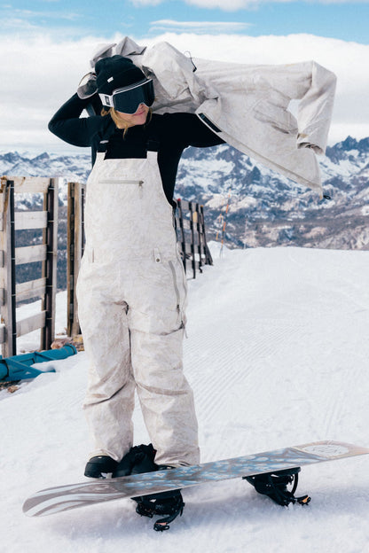 Wearing the 686 Women’s Regalia™ Bib, a person in white snow gear and goggles stands on a snowboard, adjusting their jacket with snowy mountains and a wooden fence in the background.