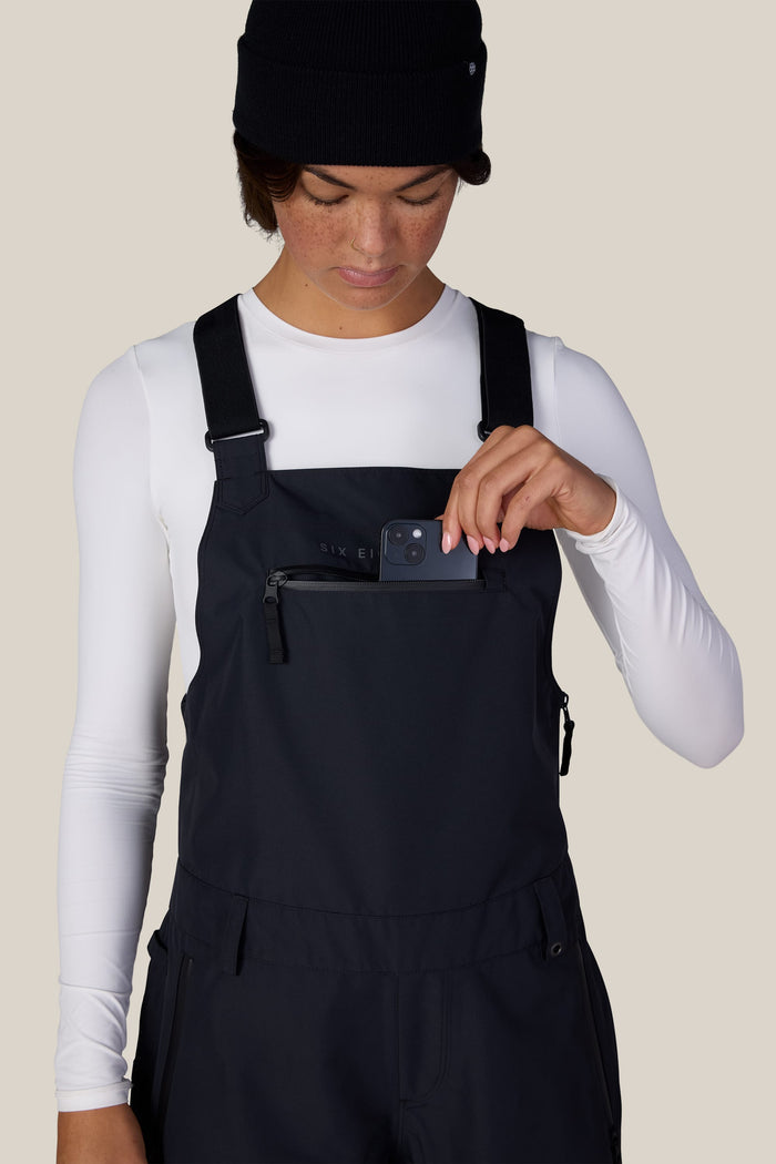 A person wearing a white long-sleeve shirt, the 686 Women’s Regalia™ Bib by 686 in black, and a black beanie places a small black device into the zippered chest pocket of the overalls, against a light beige background.