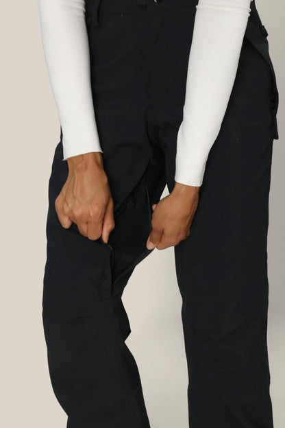 A person wearing the 686 Women’s Standard Shell Pant in black and a white long-sleeve top uses both hands to unzip a concealed zipper on the inner thigh of the pants, revealing a mesh lining beneath.