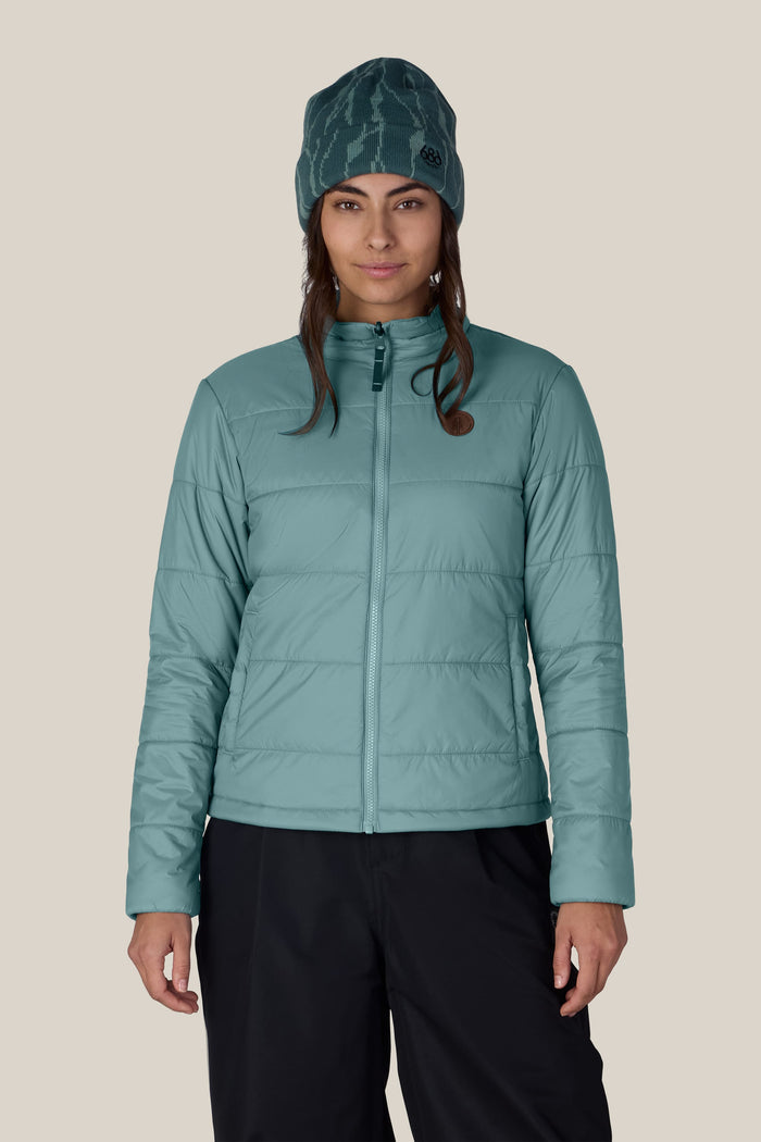 A person wearing the 686 Women’s Thermal Puff Insulated Jacket in teal, paired with dark pants and a teal beanie, stands facing forward against a plain, light background.