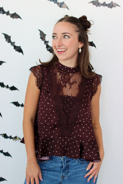 A woman wearing a sleeveless burgundy top with white polka dots and lace detailing, paired with blue jeans, standing against a white wall decorated with black bat cutouts.