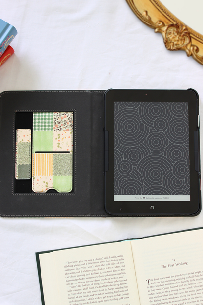 A nook case shown open on a white marble-like surface, with a patterned card holder insert on the left and a black tablet screen on the right. An open book is visible at the bottom, and part of a gold mirror frame and colorful objects appear at the edges.