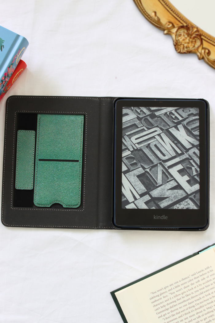 A black Kindle e-reader shown open in a folio-style case on a white fabric surface, with kickstand and hand strap. Decorative books and an ornate gold frame are partially visible around it.