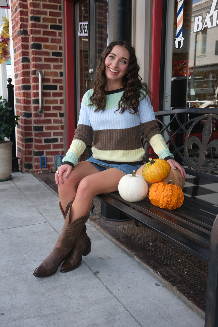 Woman wearing a long sleeve top with brown, green, blue, and yellow stripe and blue denim skirt.