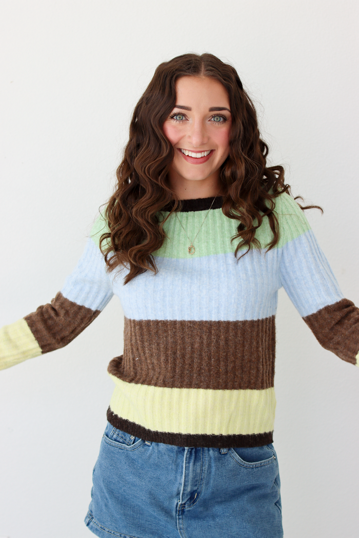 Woman wearing a long sleeve top with brown, green, blue, and yellow stripe and blue denim skirt.