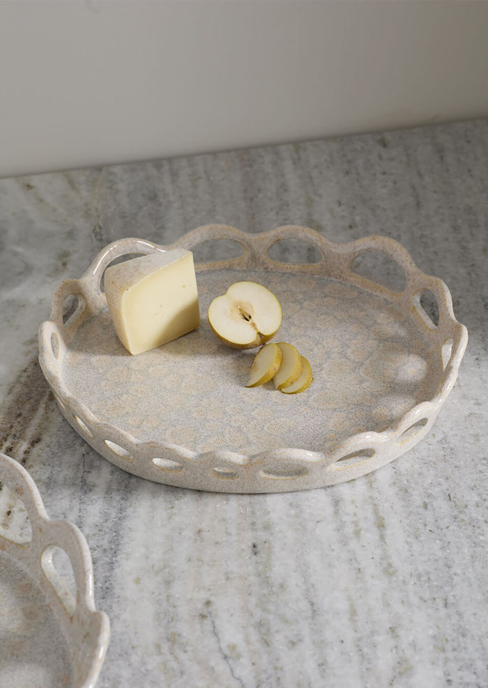 A round ceramic serving tray with a scalloped cutout edge, shown on a marble surface with cheese and apple slices as props.