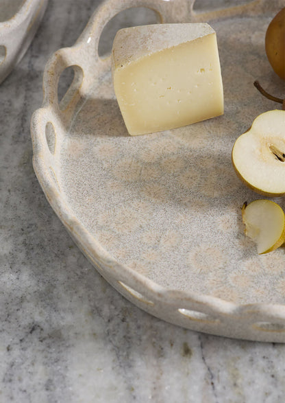 A close-up of a round speckled beige ceramic serving tray with scalloped handles, styled with a wedge of cheese, a sliced pear, and a whole pear on a gray marble surface.